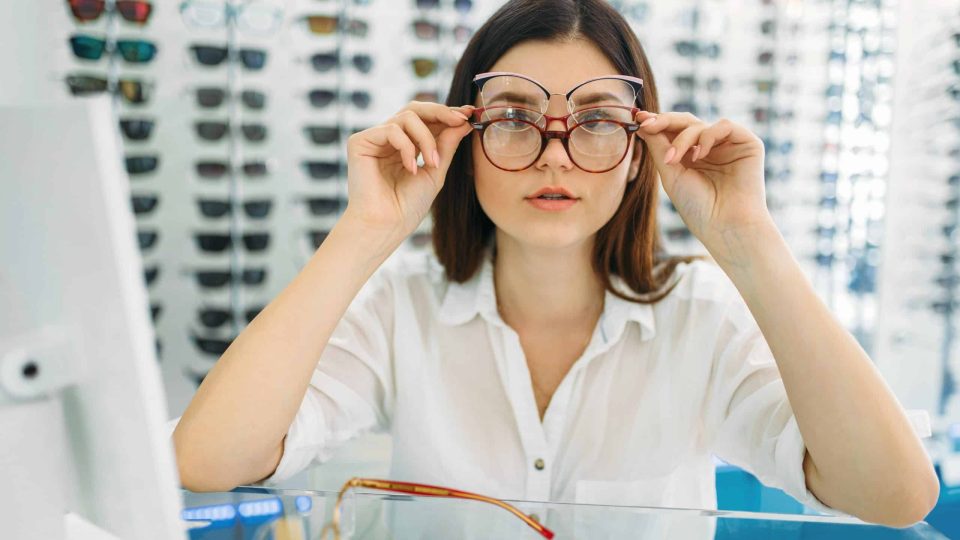 Female buyer tries on many glasses at the same time, optics store, showcase with spectacles on background. Professional eyecare, eyeglasses shop