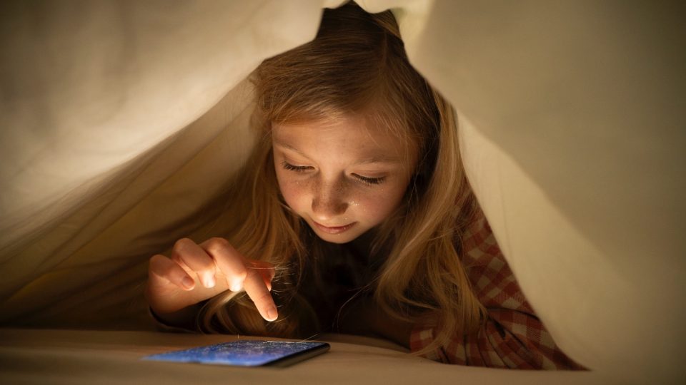 Girl using smart phone under blanket on bed at home