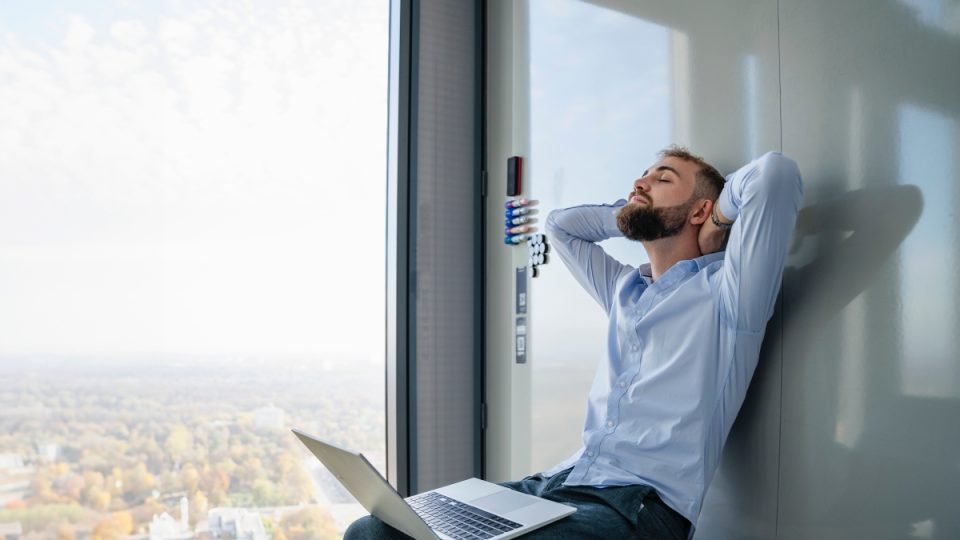 Young man relaxing with laptop in modern office with city view