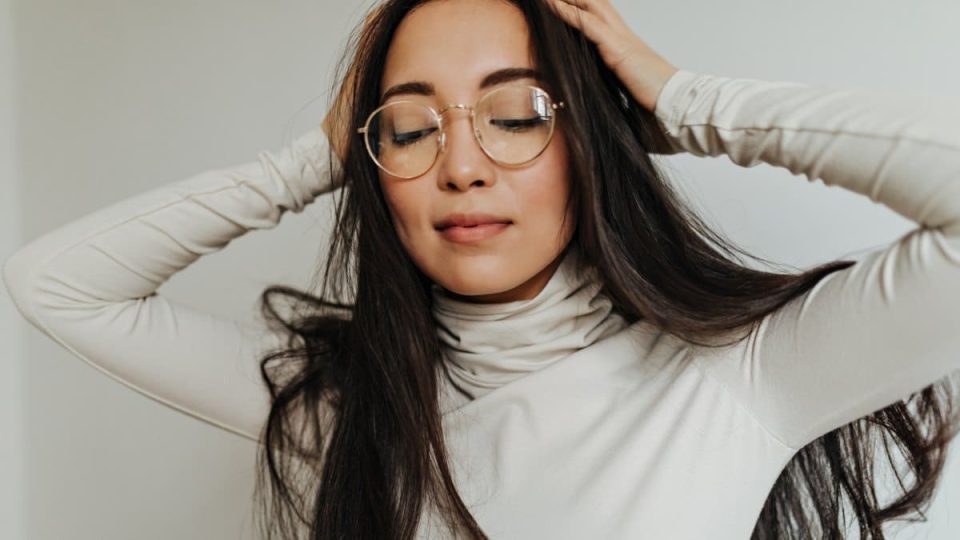 Long-haired woman in white sweater and glasses posing on isolated background and massaging her head